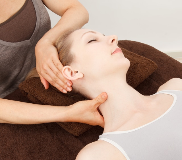 A woman receives a chiropractic adjustment while lying on her back with her neck supported by a therapist's hands.